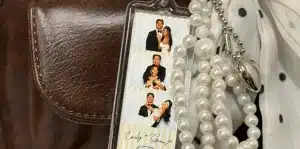 A photo booth keychain with three photo booth strips of a couple in formal attire, next to a pearl necklace and a polka-dot scarf on a brown leather surface.