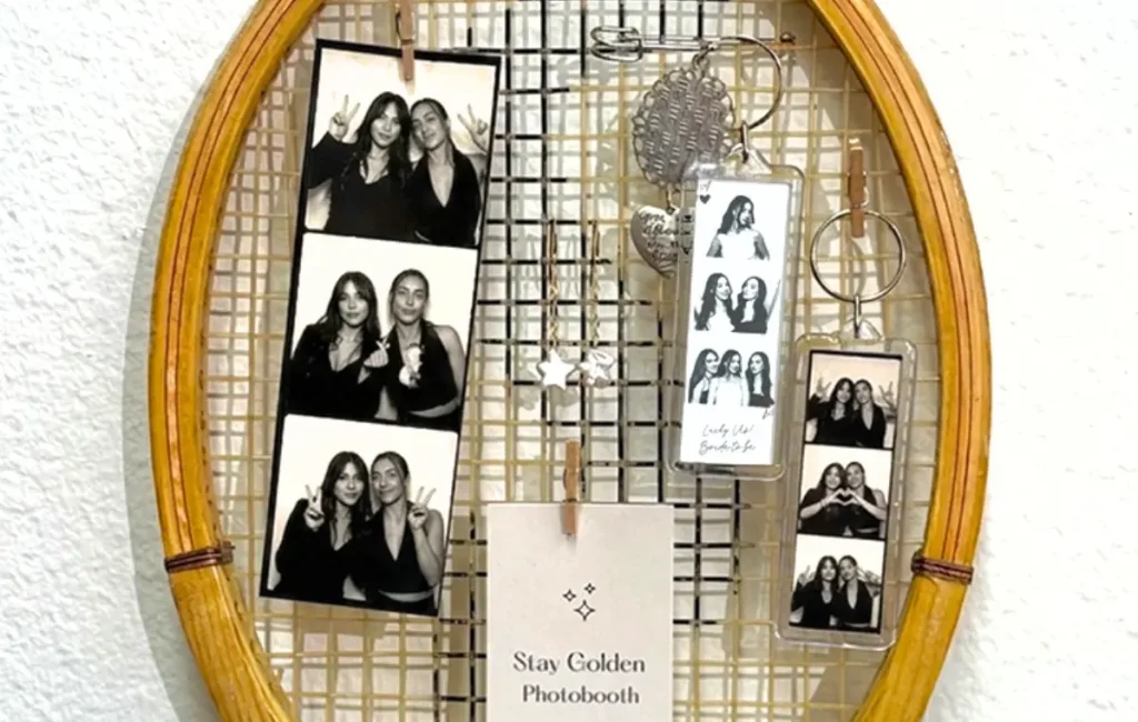 A vintage tennis racket, Event by Martina Navratilova, is decorated with photo booth keychain strips and a small card that reads "Stay Golden Photobooth."