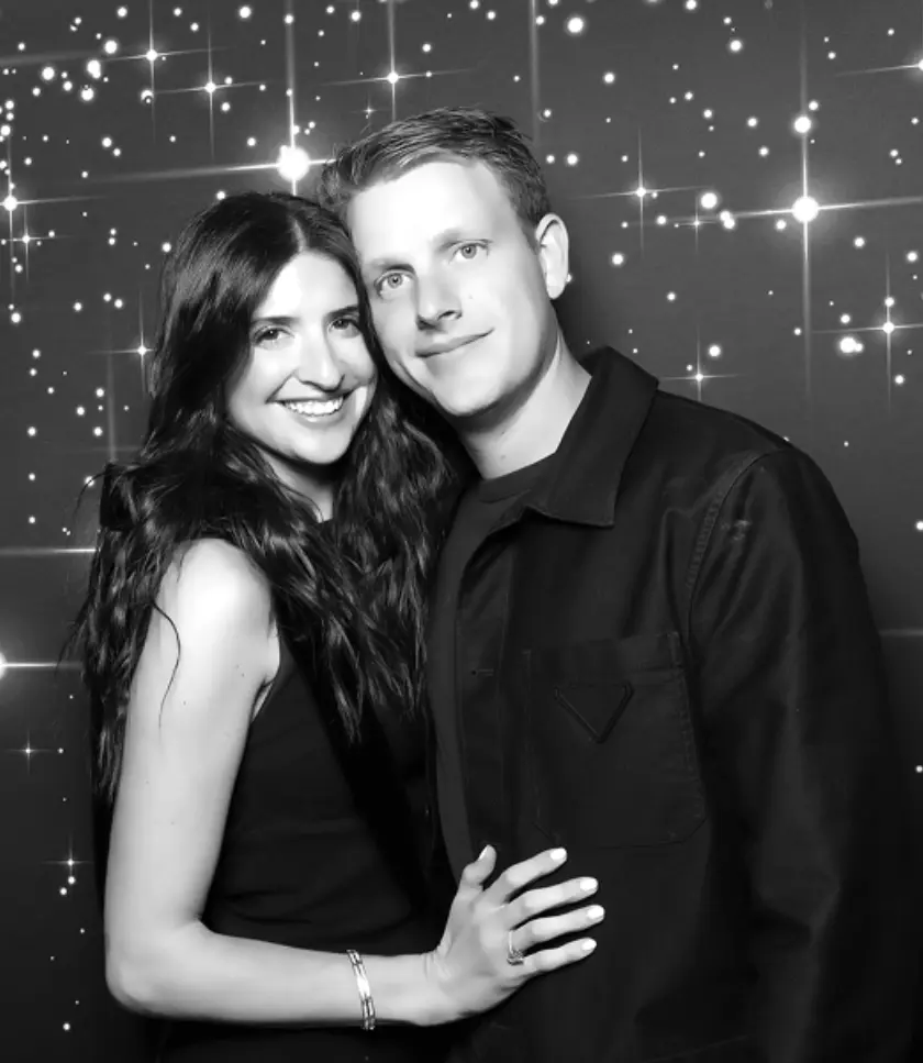 A black and white photo of a couple posing for a picture with a sparkly background. The woman is on the left, leaning her head towards the man. The man is on the right, looking towards the camera. Both are smiling.
