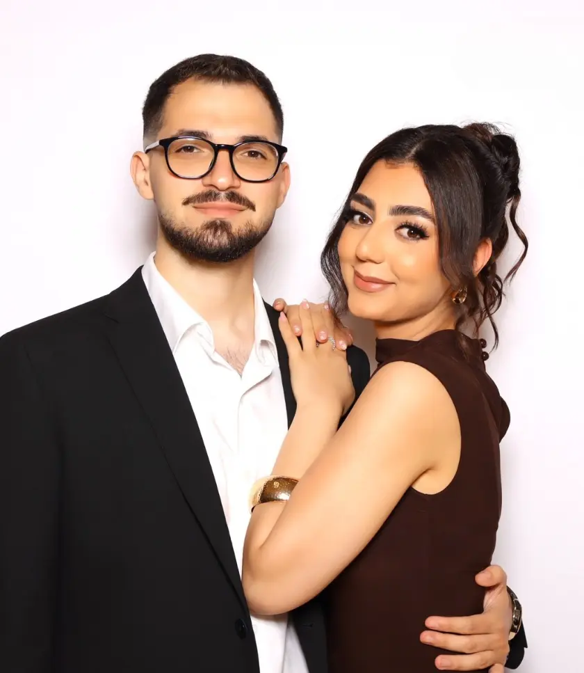 A man with glasses and a beard wearing a suit and a woman in a brown dress posing together against a white backdrop.