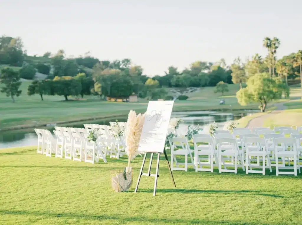 White chairs are arranged in rows on a grassy lawn facing a body of water. A welcome sign on an easel stands in the foreground, decorated with pampas grass and flowers.