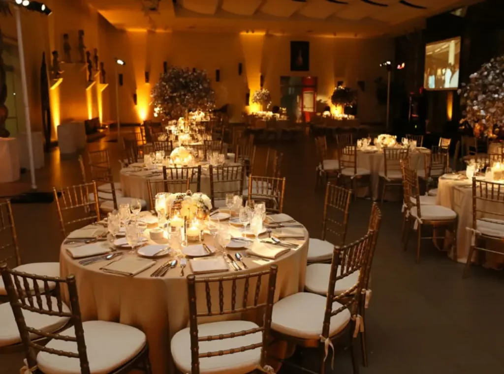 A dimly lit banquet hall is set with numerous round tables, each adorned with white tablecloths, floral centerpieces, and lit candles. Gold chairs with white cushions surround the tables, and warm ambient lighting illuminates the space.