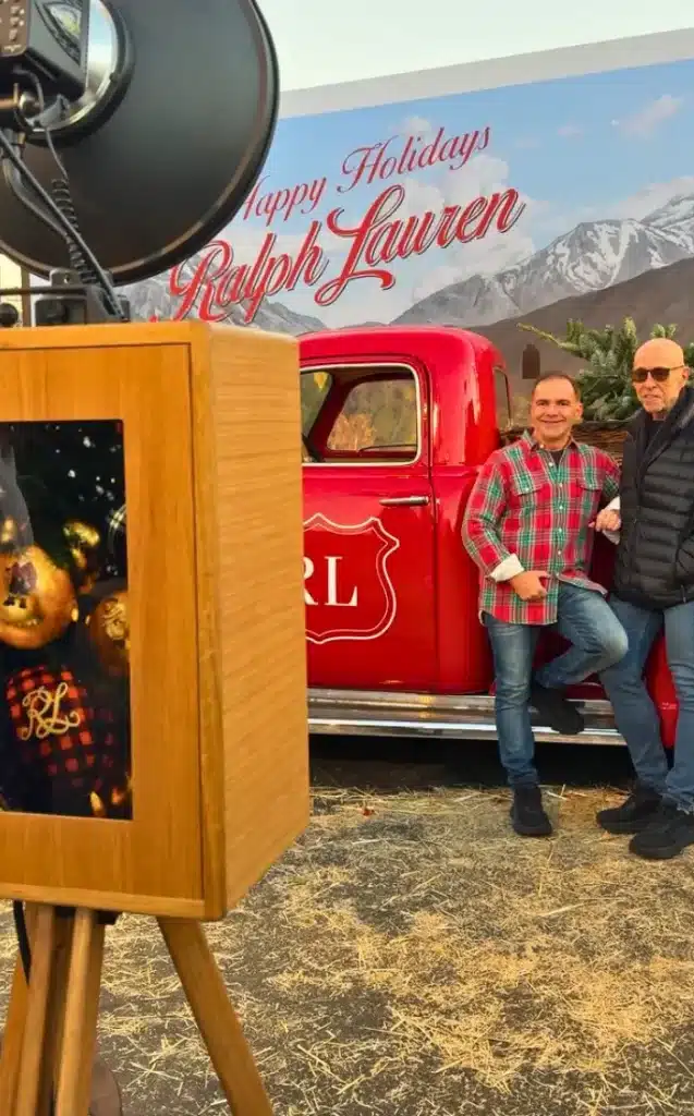 A wooden photo booth stands in front of a red truck with "Happy Holidays Ralph Lauren" written on a backdrop. Two people stand by the truck, one wearing a plaid shirt and the other a puffer jacket.