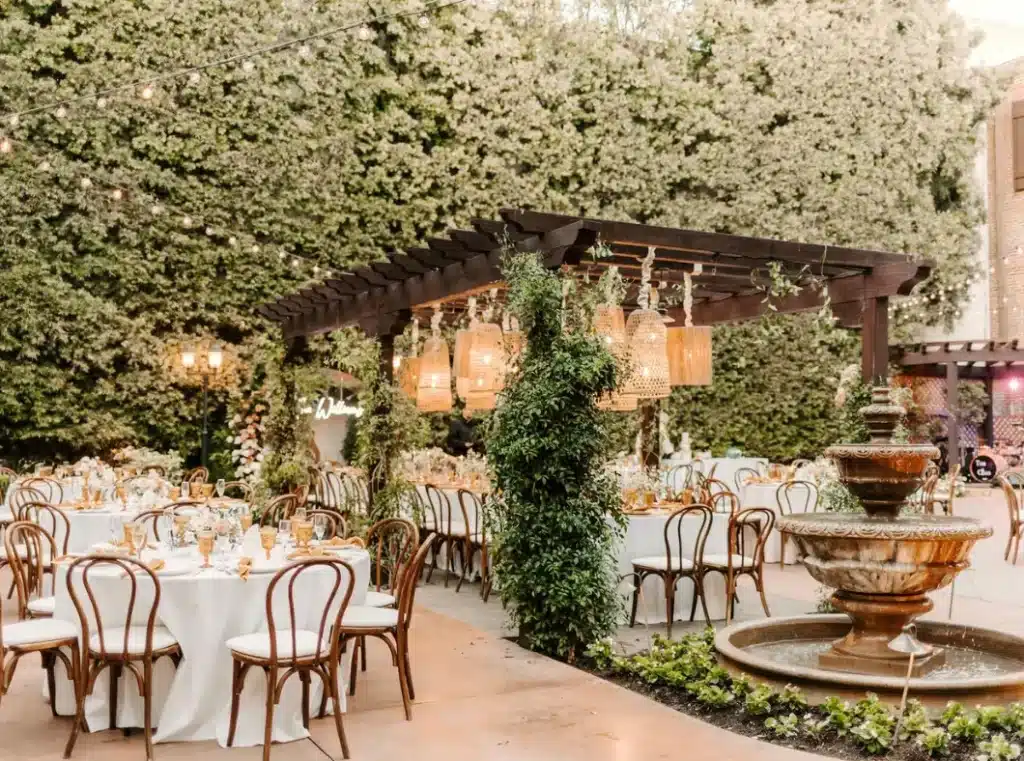An outdoor wedding reception venue with tables set for guests under a wooden pergola adorned with hanging lanterns and string lights, surrounded by lush greenery and a tiered fountain.