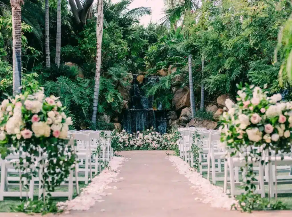An outdoor wedding aisle lined with white chairs and floral arrangements, leading towards a waterfall surrounded by lush greenery.