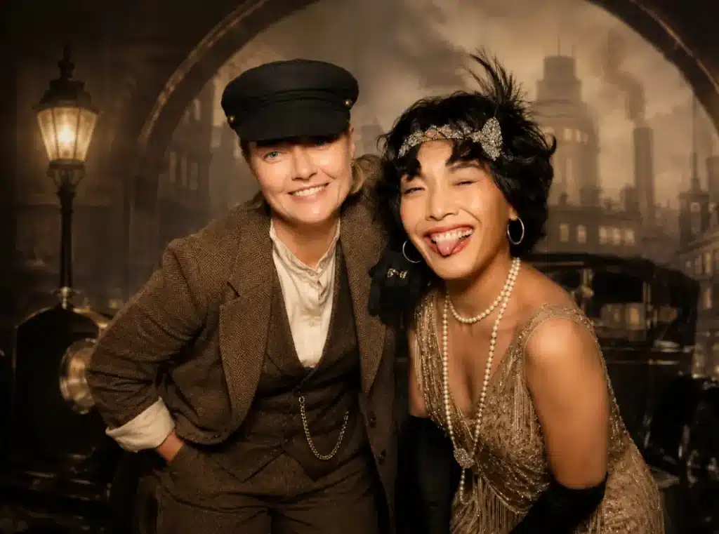 Two people posing in vintage attire against a backdrop of a city street with a vintage car and lamppost.