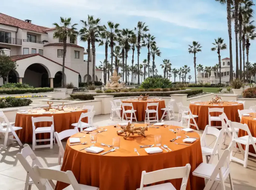 An outdoor event space with round tables covered in orange tablecloths, set with white chairs, tableware, and centerpieces, in front of a building with palm trees and a fountain.