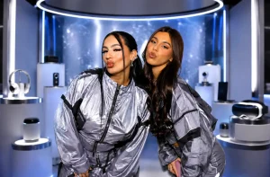 Two individuals in matching silver tracksuits pose with puckered lips in front of a futuristic display of electronics, including headphones and VR goggles.