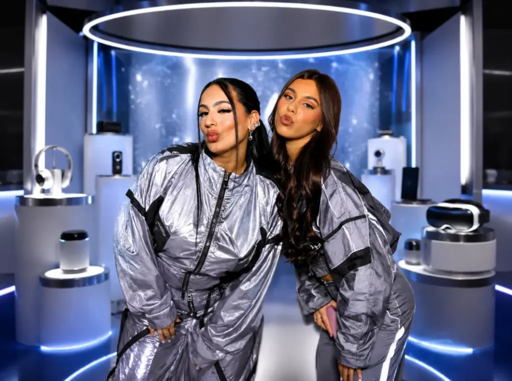 Two individuals in matching silver tracksuits pose with puckered lips in front of a futuristic display of electronics, including headphones and VR goggles.