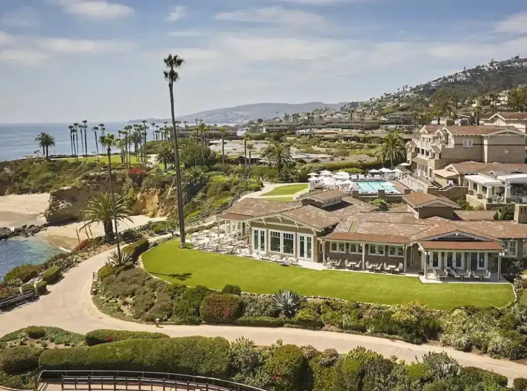 A sprawling coastal resort with Mediterranean-style architecture, lush green lawns, and palm trees overlooking the ocean.