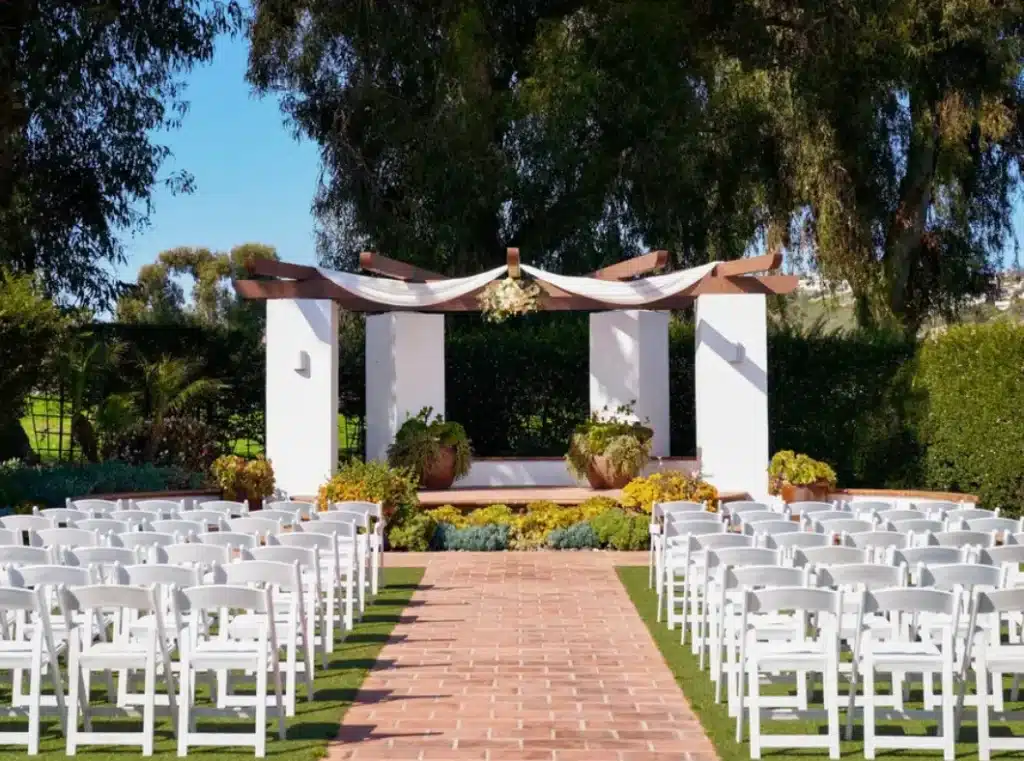 An outdoor wedding ceremony setup with rows of white chairs facing a decorated archway and altar, with a brick pathway leading to it.