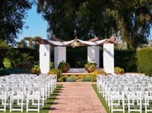 An outdoor wedding ceremony setup with rows of white chairs facing a decorated archway and altar, with a brick pathway leading to it.