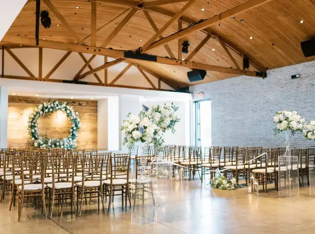 A wedding ceremony setup with rows of wooden chairs facing a floral wreath on a wooden wall. Tall floral arrangements and lanterns decorate the aisle.