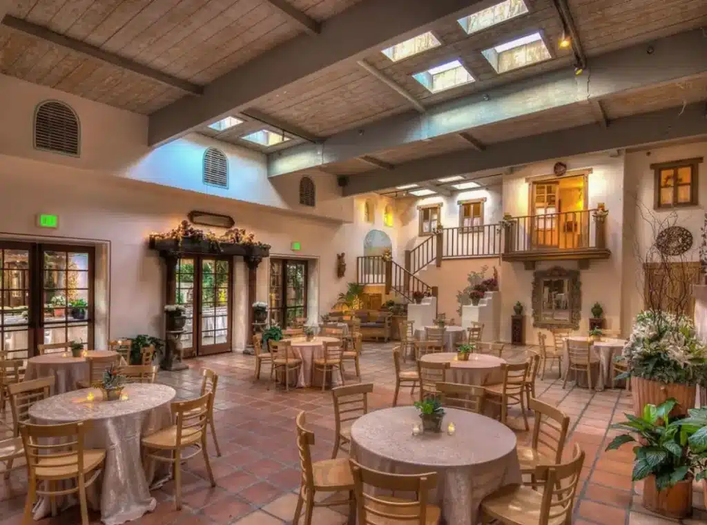 A spacious interior with terracotta tile floors, round tables set for an event, and a rustic Spanish-inspired architectural style with wooden beams and a skylight.