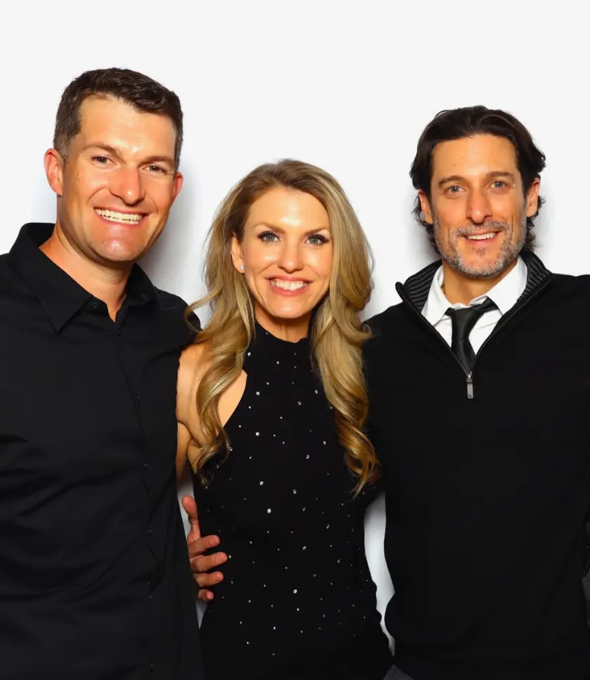 Three people posing for a photo. The person on the left wears a black collared shirt. The person in the middle wears a black sparkly dress. The person on the right wears a black zip-up sweater over a white collared shirt and tie.