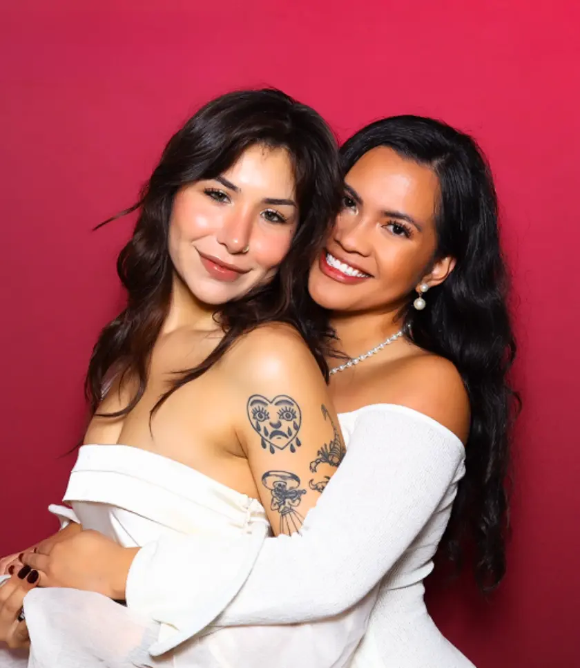 Two individuals embrace in front of a red backdrop. One wears a white off-the-shoulder dress with beaded details, and the other wears a white draped top. Tattoos are visible on the arm of the individual in the draped top.