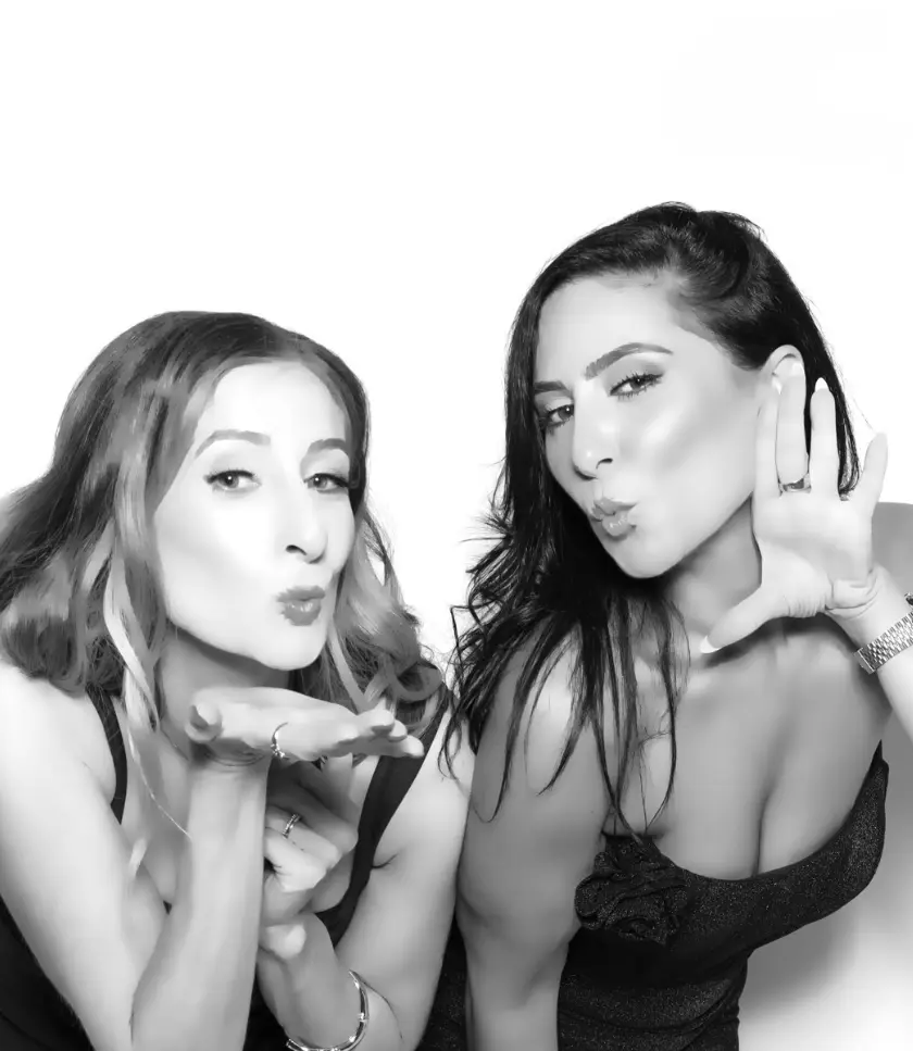 Two individuals are posing for a photo booth picture, one blowing a kiss and the other making a playful gesture with her hand.