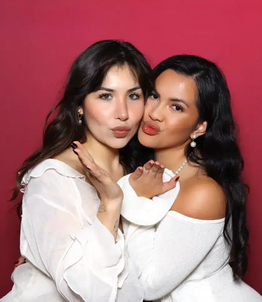 Two people in white tops pucker their lips and blow kisses against a red backdrop.