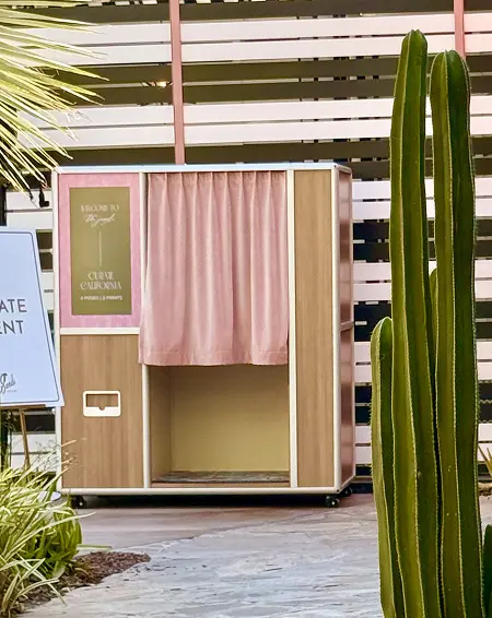 A wooden-style enclosed photo booth with a pink curtain set up outdoors beside a tall cactus.