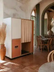 An enclosed photo booth with peach curtains stands in a sunlit room with terracotta floors, rattan chairs, and arched windows.