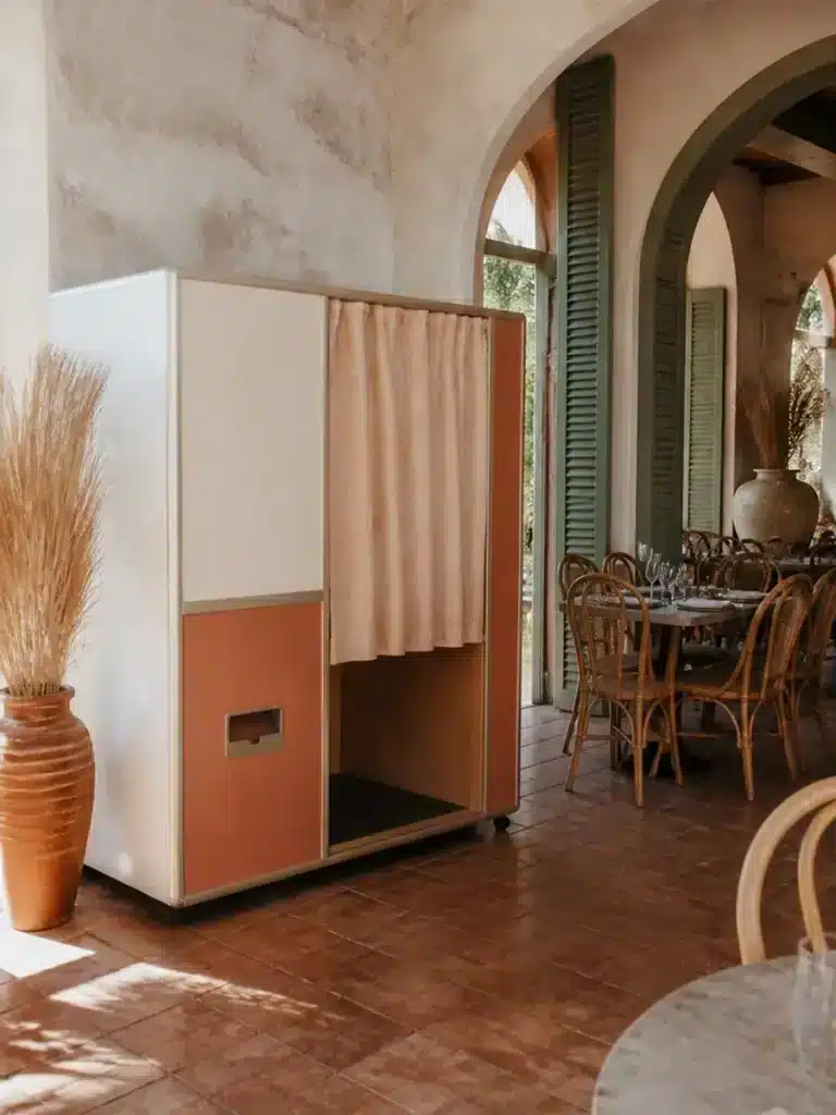 An enclosed photo booth with peach curtains stands in a sunlit room with terracotta floors, rattan chairs, and arched windows.
