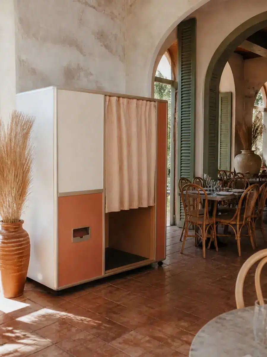 An enclosed photo booth with peach curtains stands in a sunlit room with terracotta floors, rattan chairs, and arched windows.