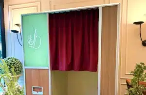 An enclosed booth with a red curtain and a green panel on the left, featuring a line drawing of a stylized figure; black-and-white checkered floor visible, surrounded by potted flowers and greenery.