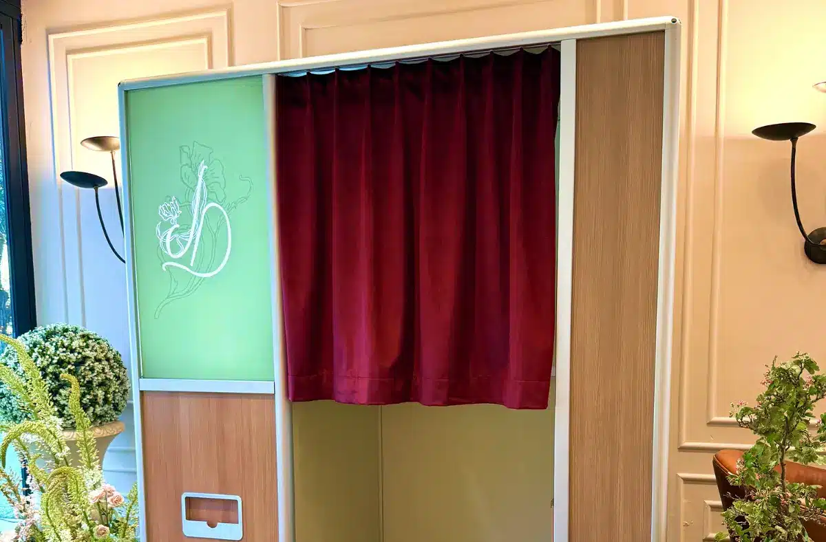 An enclosed booth with a red curtain and a green panel on the left, featuring a line drawing of a stylized figure; black-and-white checkered floor visible, surrounded by potted flowers and greenery.