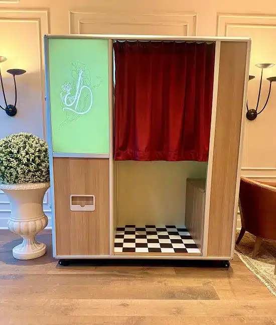 An enclosed photo booth for JD with a green panel showing minimal initials, a red curtain in the center, and a black-and-white checkerboard floor at the bottom,