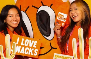 Two people pose with a large orange mascot and signs that say "I LOVE SNACKS" and "END EMPTY SNACKS".