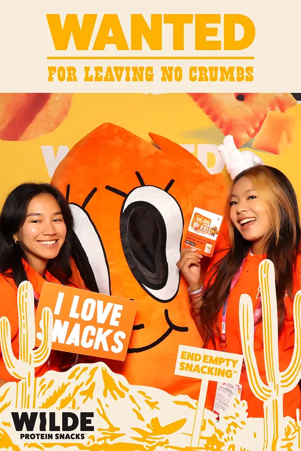 Two people pose with a large orange mascot while holding signs that read “I LOVE SNACKS” and “END EMPTY SNACKS.”
