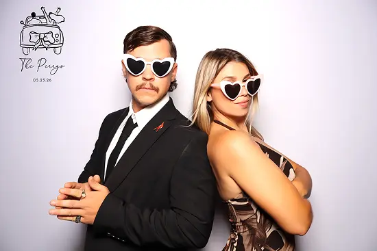 Man and woman wear heart-shaped sunglasses and pose back-to-back with crossed arms.