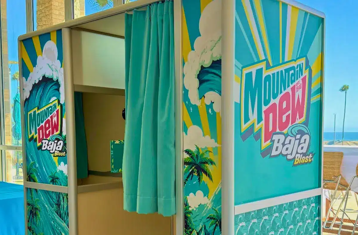 A storefront with teal and yellow beach-themed banners reading “Mountain Dew,” featuring palm tree and wave graphics, with green curtains hanging inside.