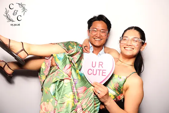 Smiling couple playfully poses with a heart-shaped “U R CUTE” sign in a photo booth.