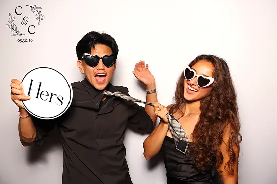 Couple wears heart sunglasses and playfully poses with a “Hers” sign and necktie in a photo booth.