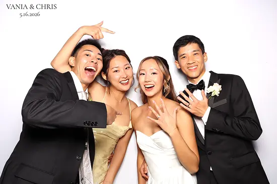 Bride and groom pose with two friends, smiling and showing off their rings in a fun photo booth pose.
