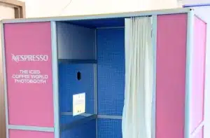 Pink and blue Nespresso booth with the text “THE ICED COFFEE WORLD PHOTOBOOTH” on the left panel, open on a blue tiled interior with a white curtain and a small device on the back wall.