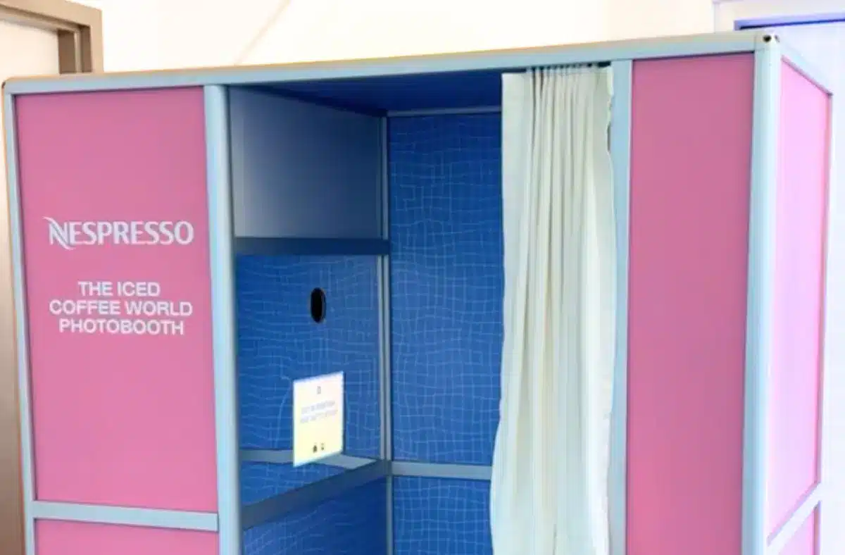 Pink and blue Nespresso booth with the text “THE ICED COFFEE WORLD PHOTOBOOTH” on the left panel, open on a blue tiled interior with a white curtain and a small device on the back wall.