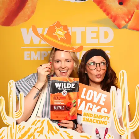 Two guests posing at a Wilde Brands photo booth, holding Wilde Protein Chips and a Love Jacks sign while wearing a cowboy hat against a bright orange branded backdrop with cactus decorations.