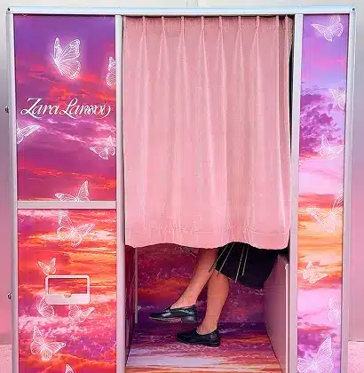 Custom Enclosed Vintage Photo Booth for Apple Music Access with Zara Larsson, featuring pink curtains, colorful sunset graphics, butterfly designs, and a guest posing inside.