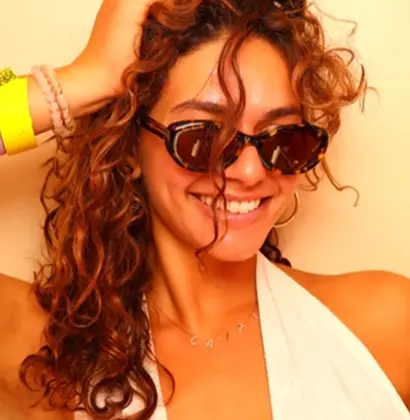Close-up portrait of a smiling guest with curly brown hair, wearing black sunglasses and a white halter top while posing inside an enclosed vintage photo booth.