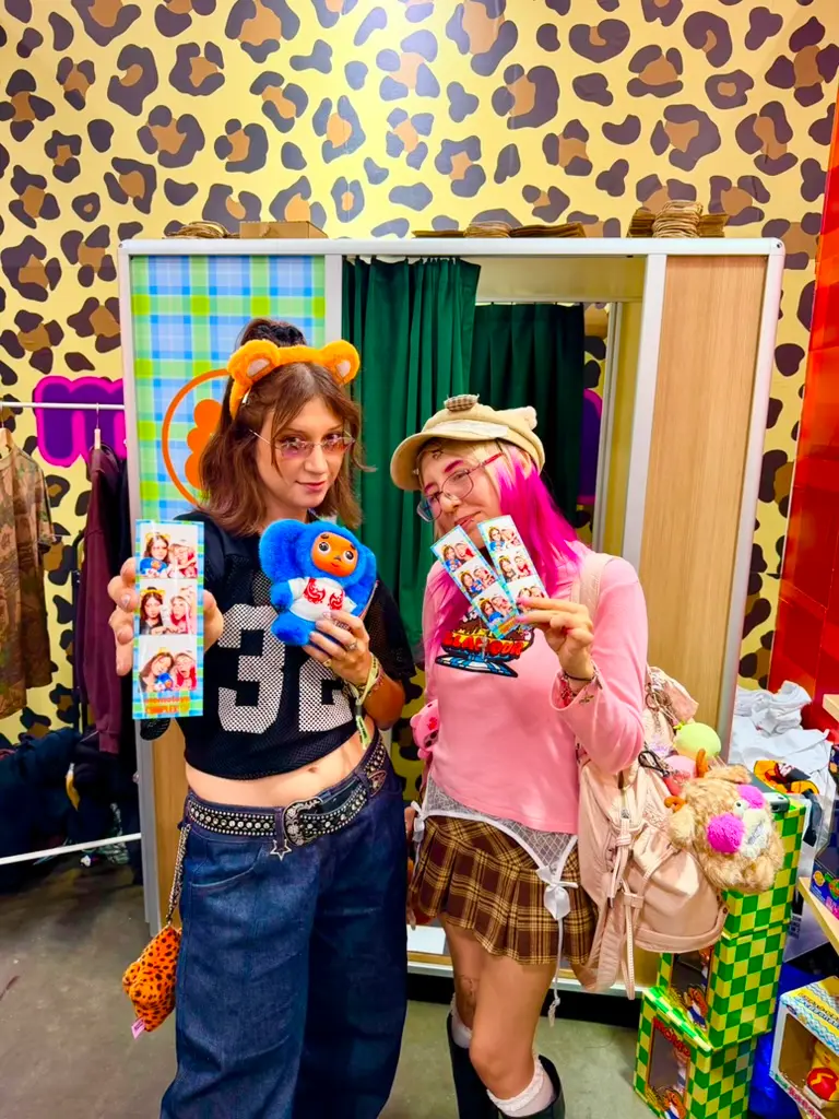Two Momotoys guests display colorful photo strips and a blue plush toy in front of a custom enclosed photo booth.