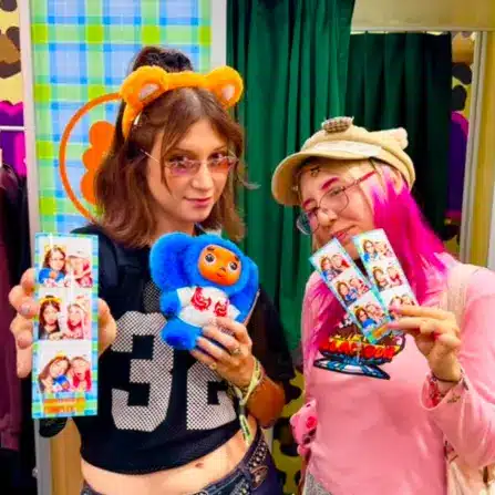 Two guests at a Momotoys event wearing playful hats and glasses, holding colorful custom photo booth strips and a blue plush toy in front of green curtains.